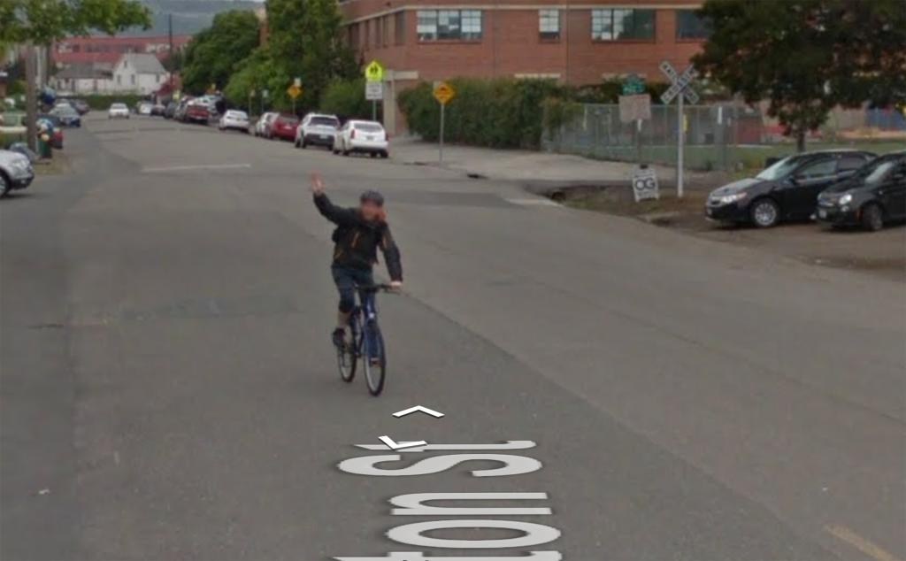 Screentshot of Google StreetView showing a street scene. On the street is a blurry figure, riding a bike and waving at the camera.